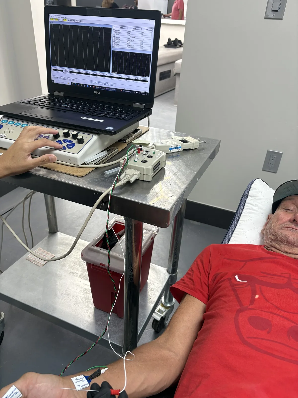 Technician operating the EMG recording console while a patient rests with nerve conduction electrodes taped to the forearm and hand at Padda Institute, St. Louis