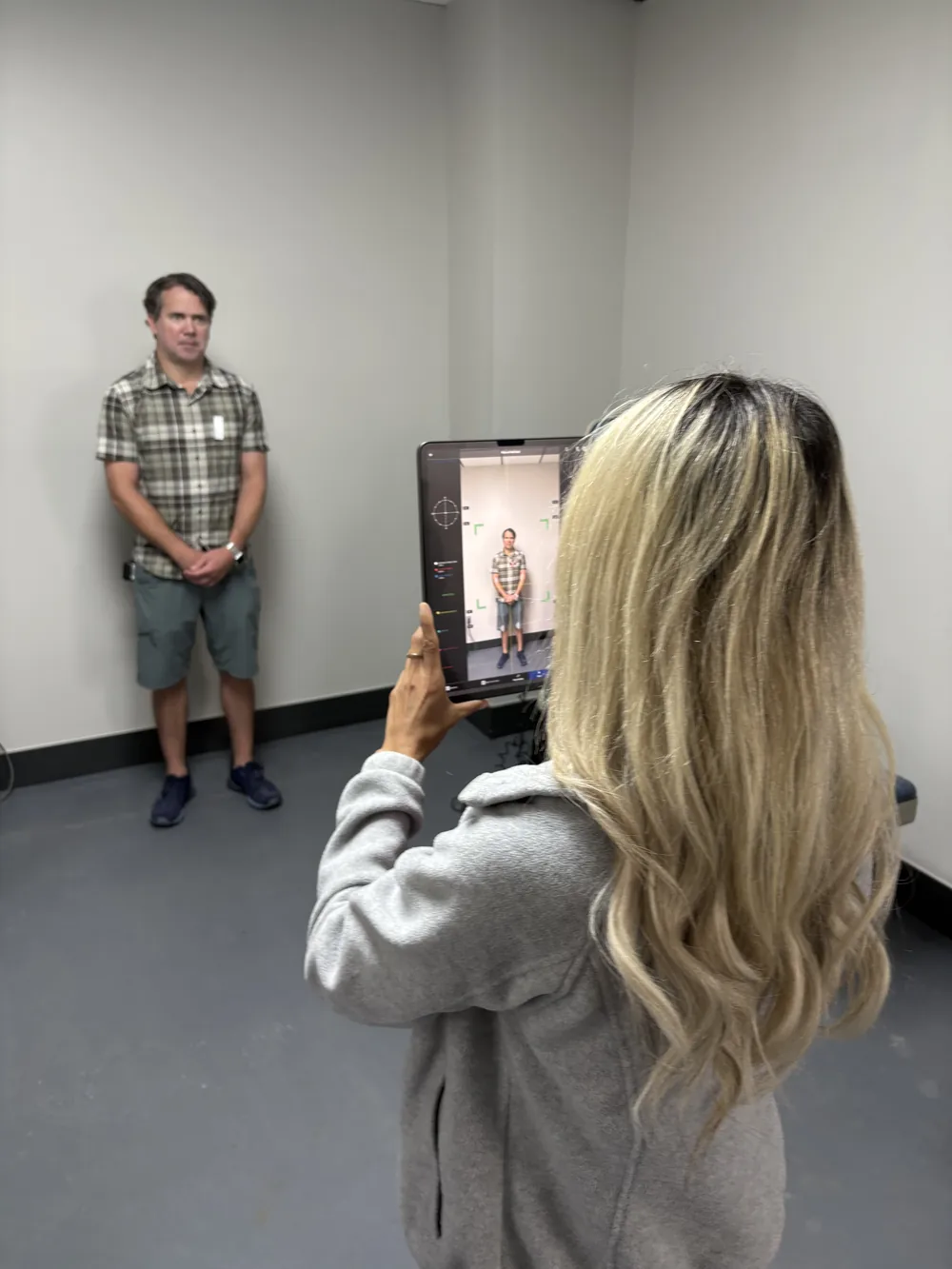 Clinician holding a tablet running Kinetisense [Gait Analysis for Fall Analysis] markerless motion capture with joint tracking markers and center-of-mass crosshairs overlaid on a patient standing for postural and gait assessment at Padda Institute, St. Louis