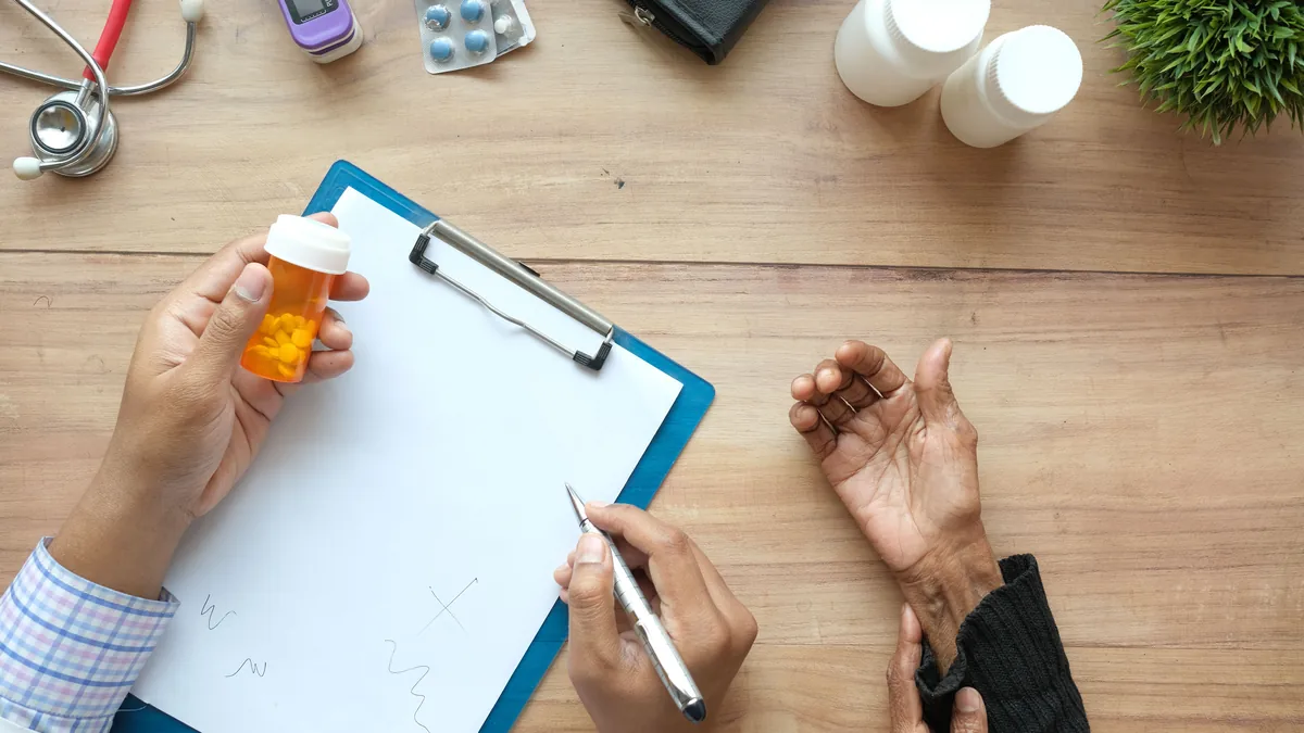 A clinician holding a prescription bottle and writing on a clipboard while talking with a patient across a desk.