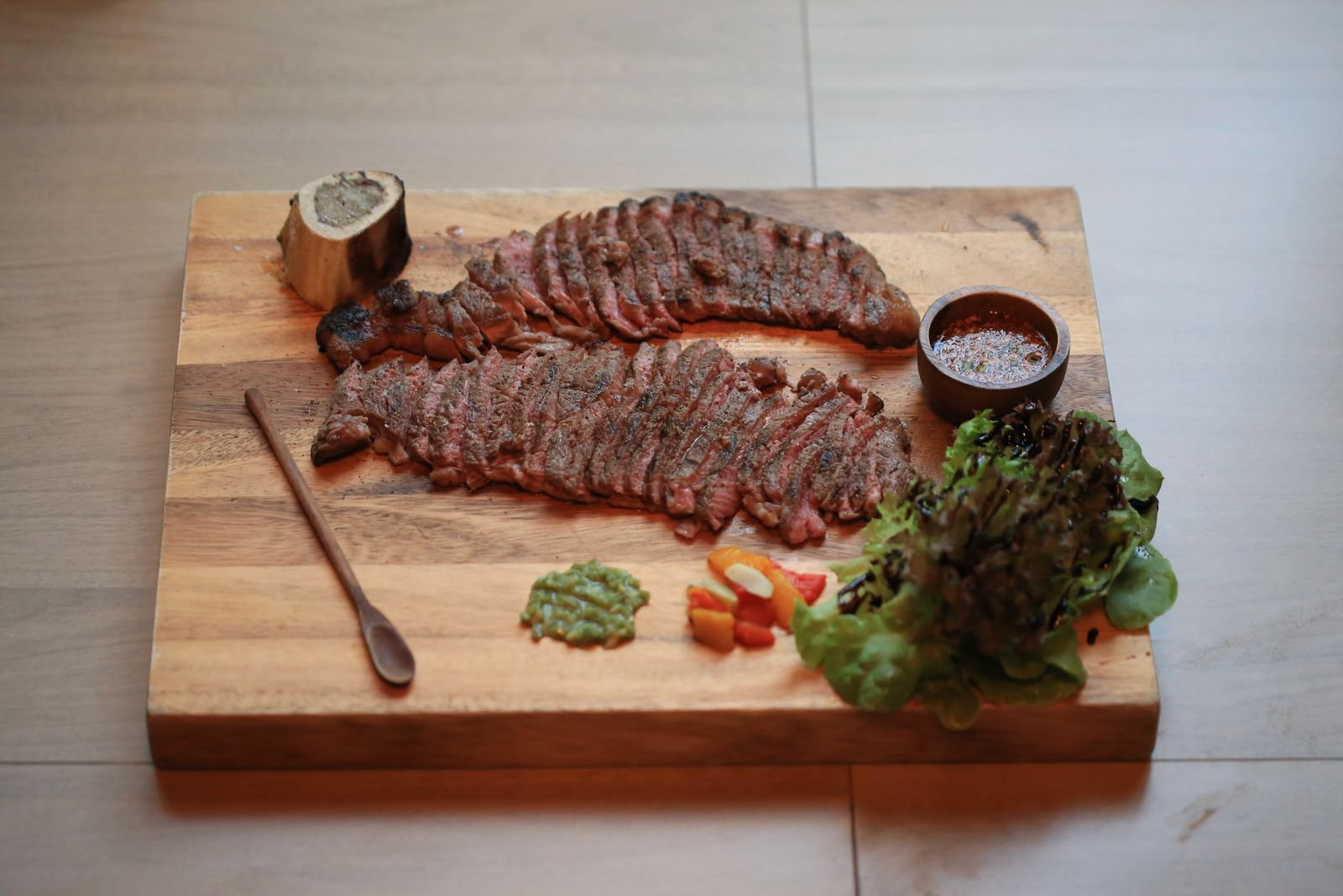 Sliced grilled steak with roasted bone marrow, green salsa, diced peppers and leafy greens on a wooden board